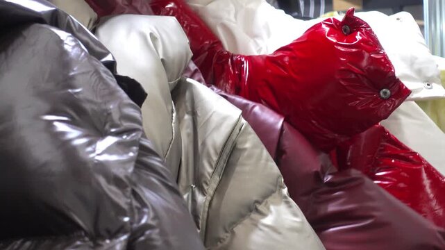 A closeup shows glossy padded winter jackets in red, silver, white, and brown as a hand browses through the rack, highlighting outerwear texture, fashion retail, and shopping