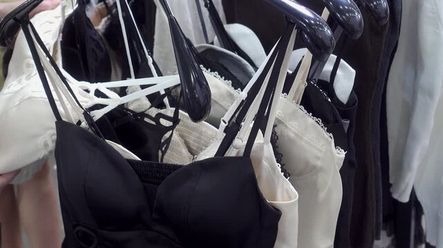Close view of hands sorting through black and white bras and delicate tops on hangers while shopping at a lingerie section inside a clothing store