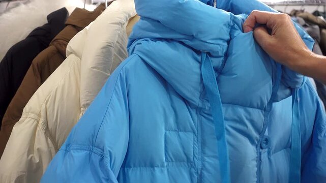 Close view of a hand checking a blue hooded puffer coat on a rack with other winter jackets in a clothing store, highlighting outerwear selection and shopping