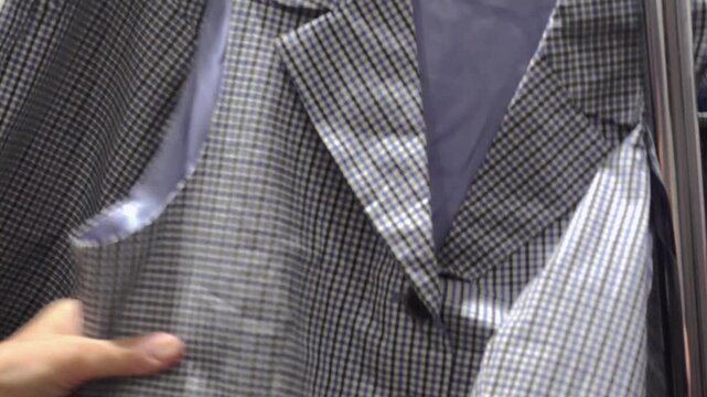 Close view of a shopper touching and examining a blue checked blazer with lace cuffs and pocket detail while browsing garments on hangers in a retail clothing store
