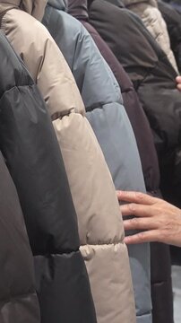 Close view of a shopper touching and browsing padded winter coats hanging on a rack inside a clothing store, comparing quilted outerwear in neutral colors