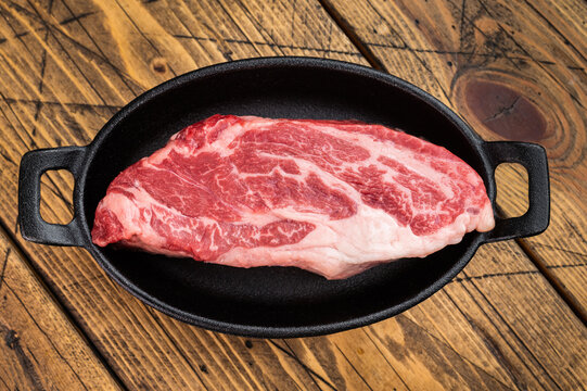 Fresh raw beef chuck eye roll cut in skillet with herbs on wood background. Beef meat ingredient overhead with copy space.