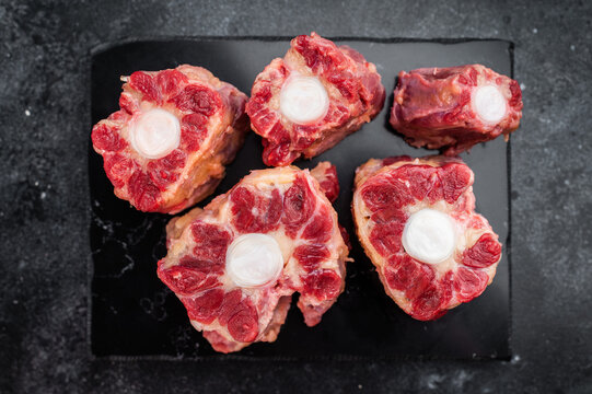 Beef oxtail sections ready for cooking on black plate, overhead composition on dramatic dark surface with space for text