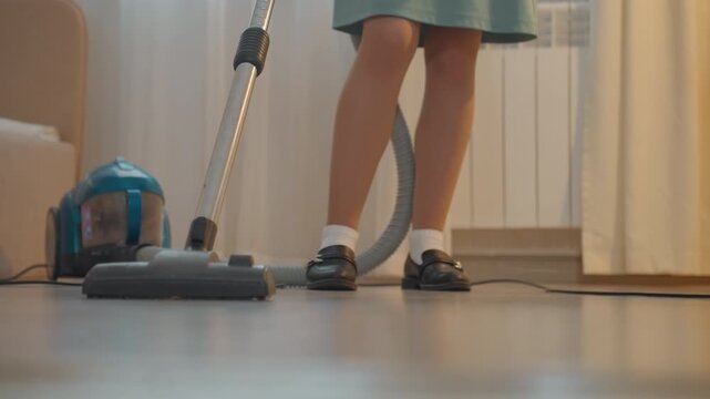 White child vacuuming living room floor, school uniform skirt, black maryjanes and white socks, blue canister vacuum gliding across polished hardwood, rhythmic backandforth strokes, warm afternoon