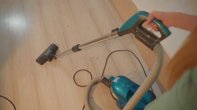 White woman vacuuming hardwood floor with blue canister vacuum and visible cord, closeup overshoulder perspective showing baseboard and dust removal, calm morning light, homeowner performing routine