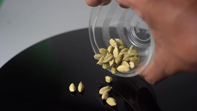 A close-up view of a hand pouring dried cardamom pods (Elettaria cardamomum) from a clear glass bowl onto a sleek, reflective black surface