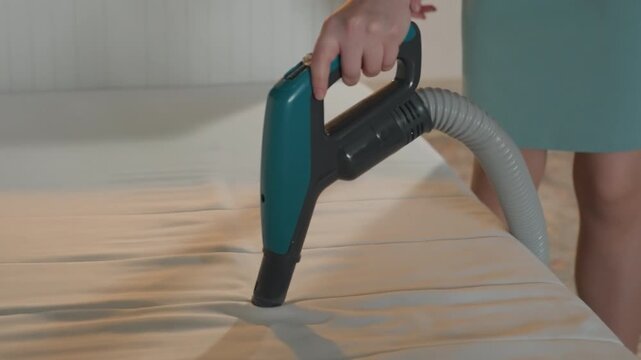 Woman vacuuming mattress in bedroom handheld extractor glides across quilted fabric closeup on nozzle suction lifting dust and debris teal dress visible calm domestic setting morning light satisfying