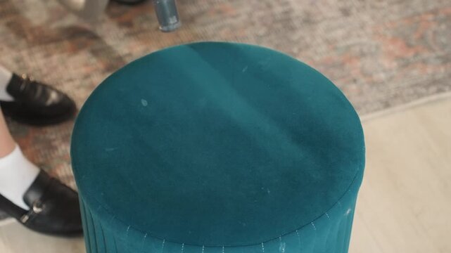 Teal ottoman vacuumed in living room beside visible shoes, handheld nozzle moves across fabric pile removing dust and debris, feet and socks indicate homeowner tending house chores, quick tidy routine