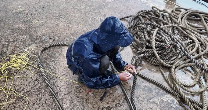 Close-up of an unidentified fisherman in a blue raincoat knotting a thick rope on the dock during a rainy day.