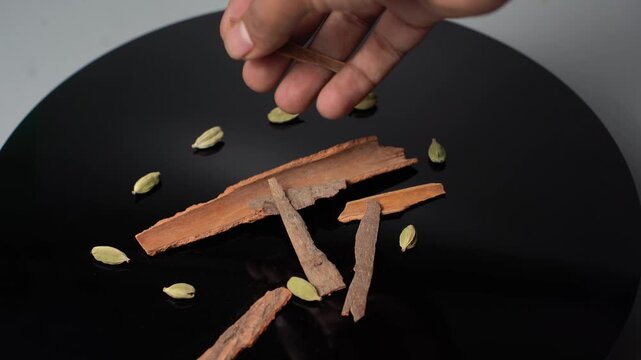 A close-up shot of a hand placing cinnamon sticks and green cardamom pods on a black plate, showcasing aromatic spices for cooking, baking, or drinks