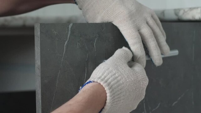 Craftsman in protective gloves fits a metallic handle to a dark kitchen cabinet door before installation. Experienced assembler holds tools and aligns hardware during home furniture renovation
