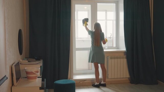 Woman cleans window. Active woman sanitizes window with cloth and spray bottle thoroughly. Woman is vigorously cleansing glass surface employing cloth and disinfectant spray bottle