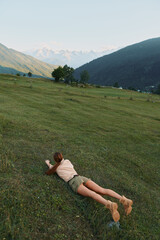 Obraz premium A young woman lies prone on a sunlit grassy hillside in a quiet valley, with distant mountains in view, conveying relaxation, freedom, and connection with nature.
