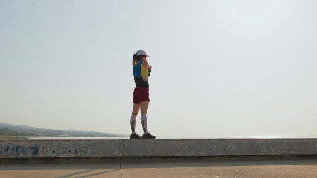 Young woman stands on a concrete pier, facing the vast ocean. She wears red shorts, white socks, and a blue helmet. Arms raised high, eyes closed, serene smile visible. Perfect for travel, adventure