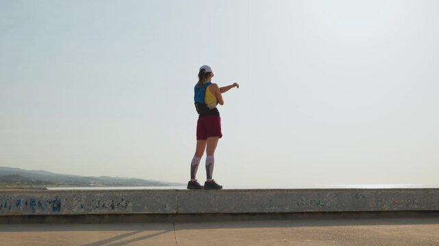 Person stands on concrete seawall, arms raised high. Back to camera, confident pose. Points toward distant ocean. Clear sky, calm sea visible. Happy traveler celebrates new view