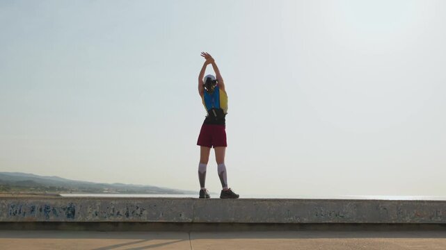 Young woman stretches on a concrete sea ledge. She wears a red skirt and blue top. Her backpack rests on her back. She faces away, arms raised high. Hazy mountains glow behind her