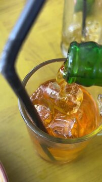 Pouring Indonesian iced jamu temulawak, traditional herbal drink with ice cubes on wooden table, slow motio
