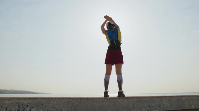Person stretches on a concrete pier at sunset. Sunlight glints off the calm sea. They hold a board and wear sportswear. Their expression shows focus and readiness. Ideal for fitness, wellness