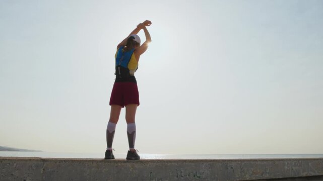 Runner stretches arms high toward sunset. Backpack visible on shoulders. Person appears relaxed and satisfied. Perfect for fitness or wellness content. Calm coastal environment