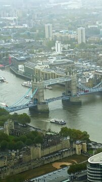 High angle drone footage capturing the iconic Tower Bridge spanning the River Thames, with the historic Tower of London on the riverbank and boats passing on a cloudy, atmospheric day