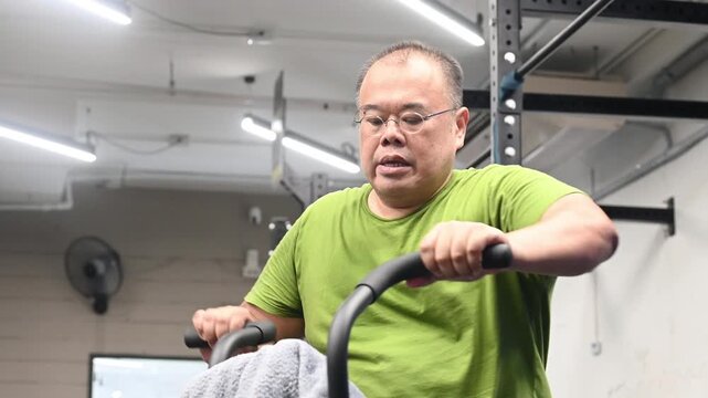 Exercise for overweight man cycling on indoor bike in gym focused and determined wearing green shirt working out with towel pedaling stationary bike with focused expression during gym workout fitness