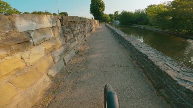 First person view of bike ride on paved path beside canal. Stone walls line both sides. Warm golden light bathes scene. Trees and greenery frame waterway. Peaceful in the park during sunset