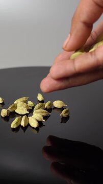 A close-up view of a hand carefully dropping green cardamom pods onto a shiny black surface, creating a small pile. The lighting highlights the texture of the spice