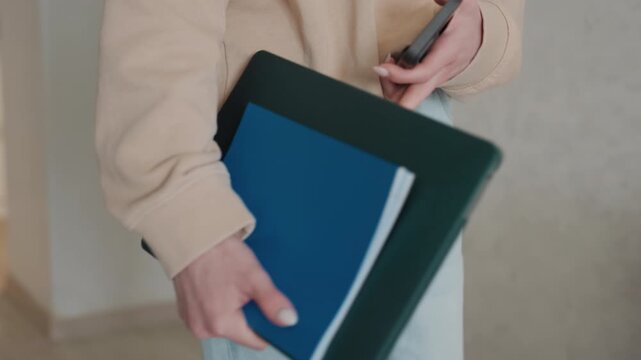 Caucasian tutor placing folders on desk in cozy home entryway, carrying keys and phone, lays blue binder and notebook beside lamp and potted plant, casual jeans and sweater, preparing grammar lesson