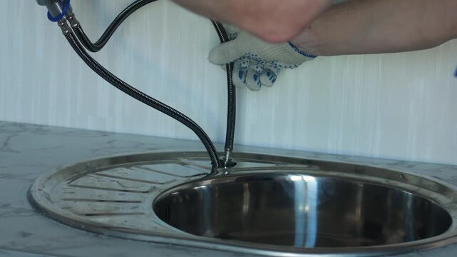 Plumber threads flexible water supply hoses through the kitchen sink hole and installs a new faucet. Experienced craftsman wearing protective gloves aligns and secures modern tap on the sink properly