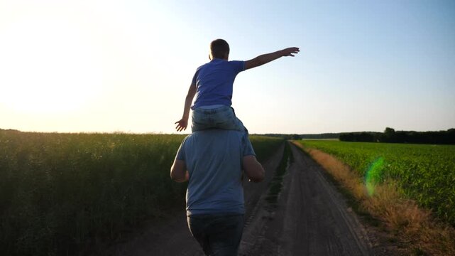 Little boy sitting on shoulders of his daddy and playing raised hands as airplane at sunset. Happy father and his son having fun at countryside. Dad with small kid spending time together outdoor