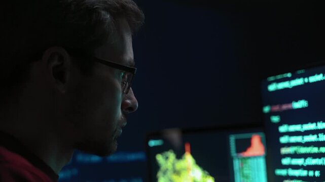 Side view close up, concentrated coder or software engineer in glasses works through a difficult debugging session at dark desk with multiple monitors late at night, lines of code running around him