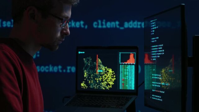 Software engineer in glasses sits at a dark desk with multiple monitors, works on data science or artificial intelligence development. Programmer writing code late at night. 