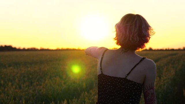 Happy female hipster with tattoos having fun during goes through green barley field at beautiful sunset. Cheerful punk girl in short black dress fooling around while walks among wheat meadow. Slow mo