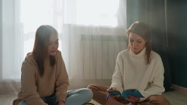 Two white women coaching exam preparation, sunlit room with notebook and planner open, pen and laptop on floor, mentor offering feedback and correction, student listening and writing, concentrated