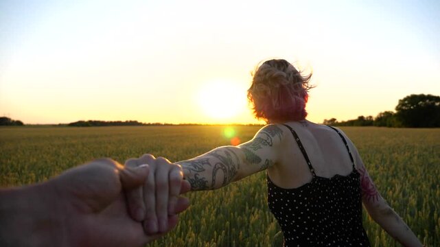 Follow me shot of joyful punk girl holds male hand jogging through barley meadow at sunset. Cheerful female hipster pulling her boyfriend among wheat field. Happy couple having fun at nature. POV