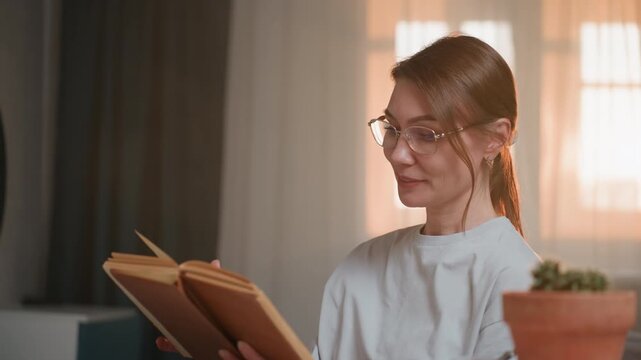 Woman reading grammar book at home, glasses and potted plant on desk, warm sunlight through curtain, closeup of calm tutor explaining rules and turning pages, soft ambient study atmosphere for online