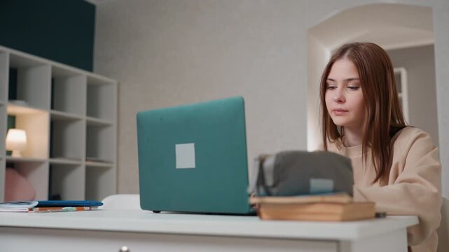 Quiet study scene woman at laptop in calm workspace with teal computer open notebook, pencil case and books on desk, soft shelf lighting, thoughtful expression during online lesson and revision