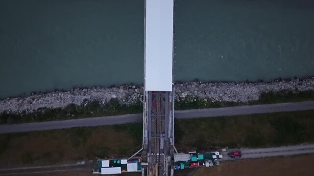 Top-down drone footage showing a railway bridge construction site with a temporary white protective roof and the interrupted railway line on both sides of the river crossing.