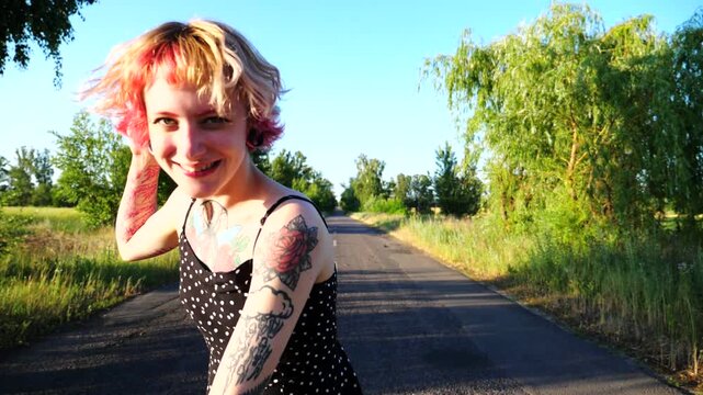 Carefree hippie woman in black dress walking along empty road at countryside. Happy punk girl with pink hair fooling around while goes on country driveway. Young female hipster having fun outdoor