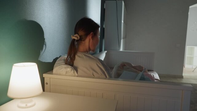 Caucasian mother soothing infant at night, tender nursery moment with bedside lamp glow, female parent holding newborn on crib edge, gentle rocking, warm soft atmosphere and quiet lullaby mood