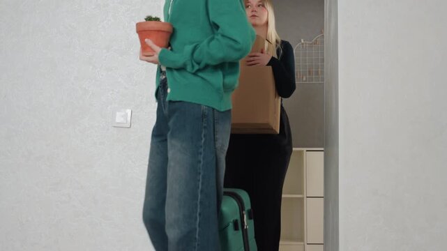 Roommates pause in doorway with boxes, one holds potted plant while other carries stacked cardboard, closeup on feet and hands, soft smiles and tired relief, grey wall and shelving backdrop, scene