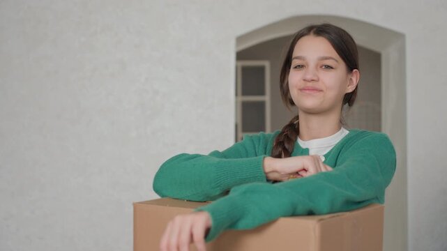 Caucasian girl holding new house keys, leaning on cardboard box with bright smile, braided hair and green sweater, upbeat apartment entry scene, firsttime homeownership emotion, playful key gesture