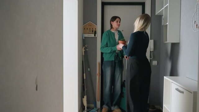 Two women carry suitcase and mug while handing keys in small foyer, cooperative move scene with green jacket, luggage on wheels, casual conversation and quick paperwork at doorway