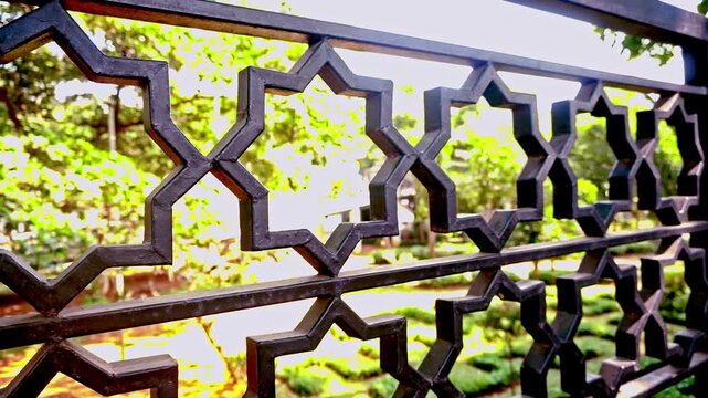 Close up shot of ornate black metal geometric railing featuring Islamic star patterns with a soft focus background of lush green trees and sunlight in a peaceful park.