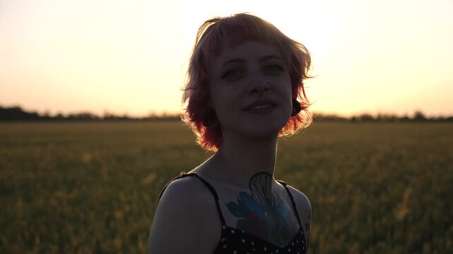 Happy punk girl with pink hair looks into camera standing on barley meadow at sunset. Portrait of smiling hippie woman with ear tunnels with wheat green field at background. Concept of freedom