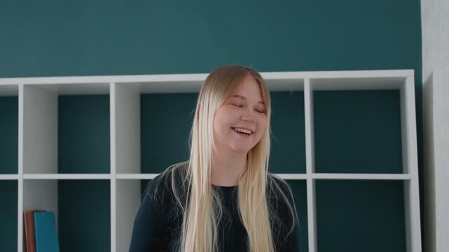 Smiling white woman in new apartment, empty shelving teal wall, blonde hair relaxed sweater, playful gestures unpacking, roommate blurred in foreground, natural daylight joyful movein moment