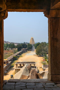 Distant view of the majestic yellow gopuram tower of Virupaksha Temple rising above lush green trees and ancient ruins along a street in Hampi, India. Historic Hindu architecture landmark.