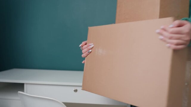 Two white women packing boxes desk new apartment with teal wall, one passing potted plant while other stacks cardboard, casual sweaters and jeans, careful hands arranging decor, minimalist workspace,