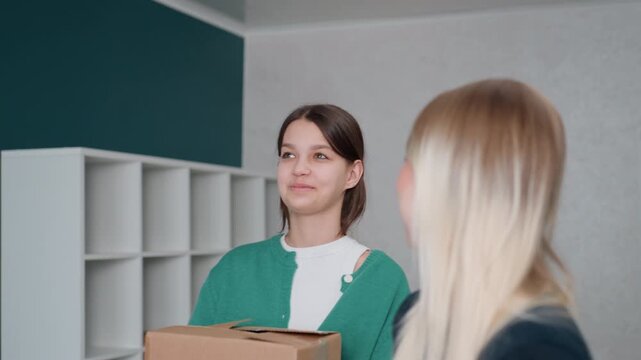 Two women carrying box inside empty apartment, teal accent wall and white cubby shelves, brunette holding cardboard while blonde watches, gentle silence and hesitant smiles, small plant on counter,