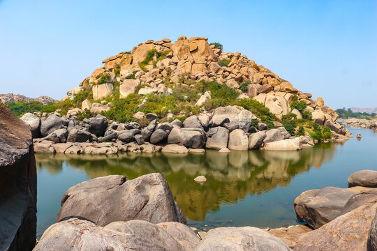 Scenic landscape of ancient rocky granite boulder hill and river with water reflection in Hampi, India. Historic nature, geological wonder and archaeological site travel destination view.